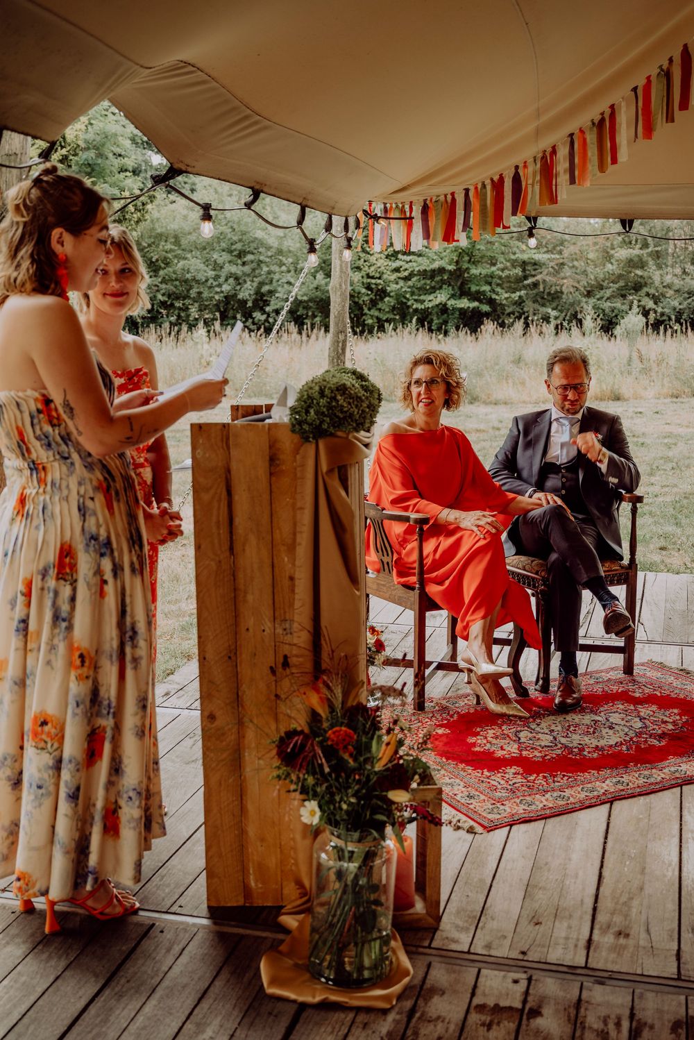 Bridesmaid in floral dress giving speech at wooden podium while couple in red and navy sit on decorated stage.