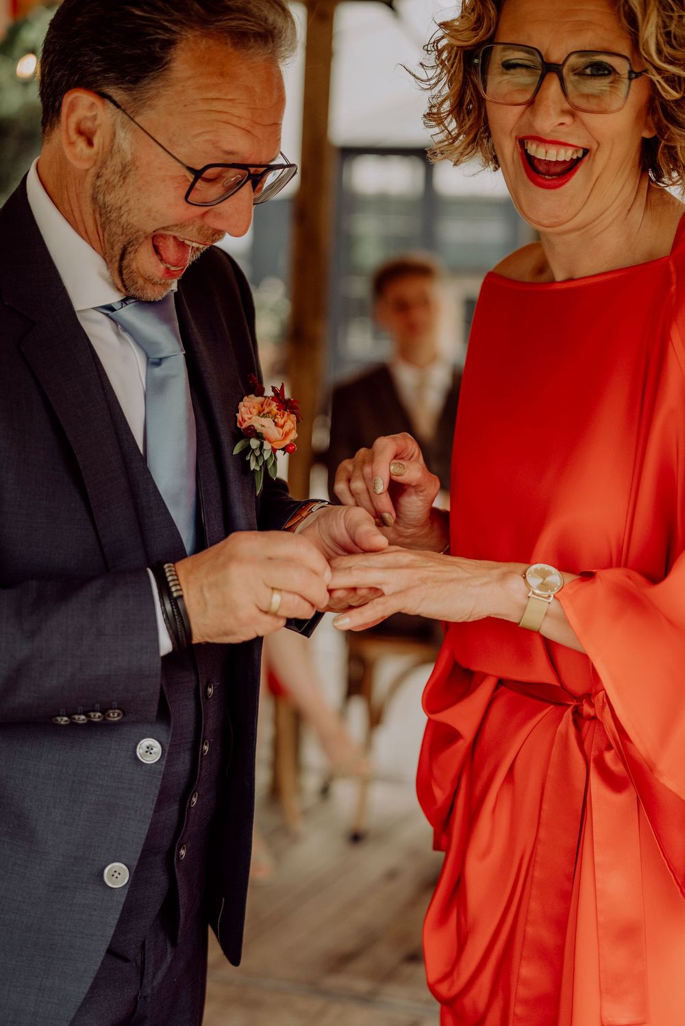Laughing bride in red dress and groom in navy suit joyfully exchanging wedding rings at ceremony.