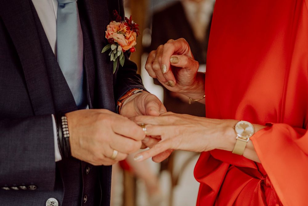 Close-up of groom placing wedding ring on bride's finger wearing red dress and silver watch.