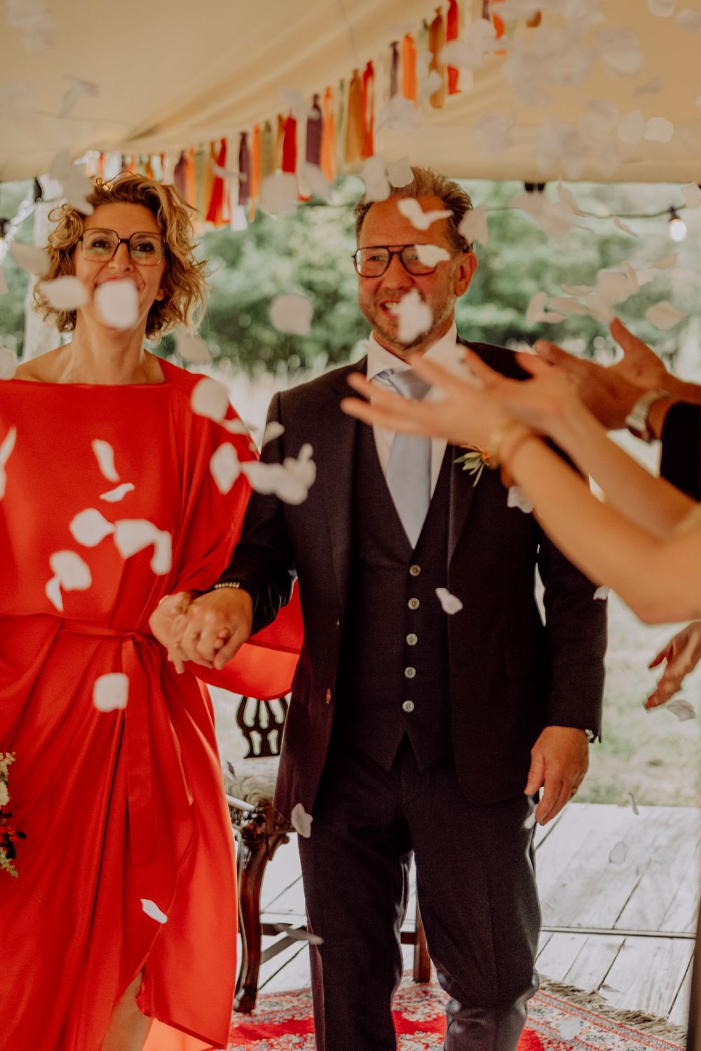 Joyful newlyweds holding hands while guests throw white flower petals under colorful tassel tent.