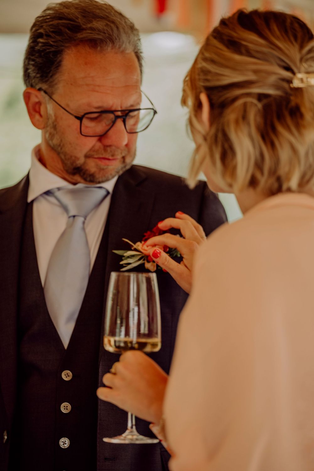 Groom in navy suit with boutonniere receiving champagne glass from guest at wedding reception.