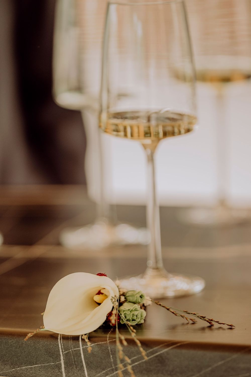 Elegant wedding boutonniere with white calla lily beside a glass of white wine on a golden table.