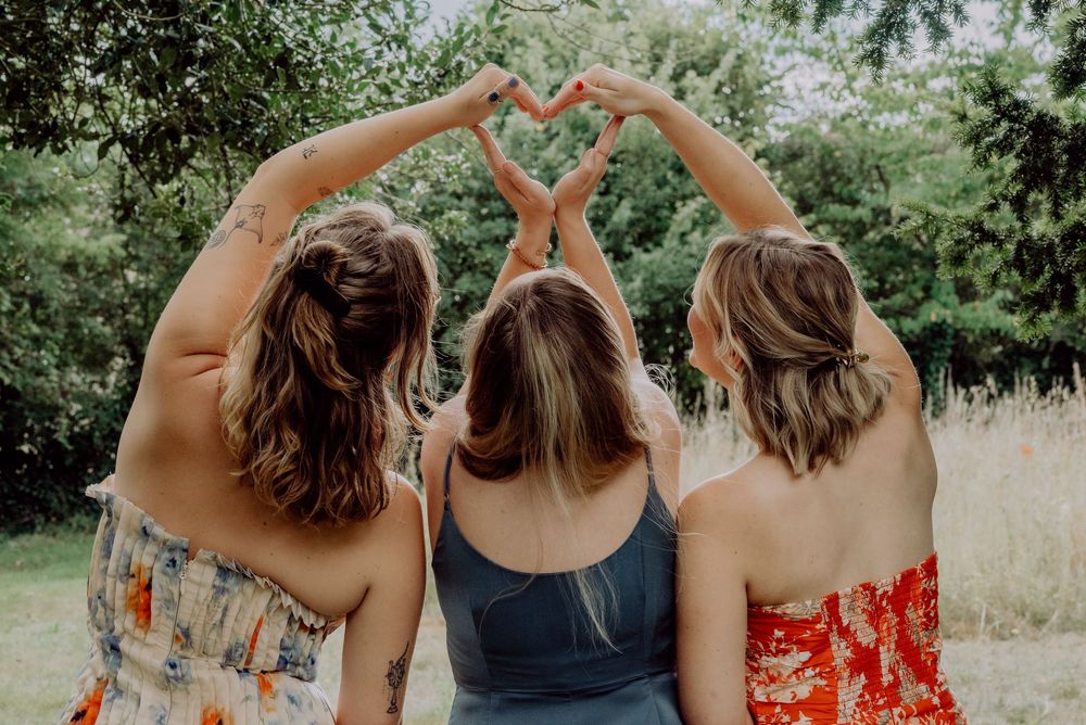 Three women sitting outdoors forming a heart shape with their hands raised above heads.