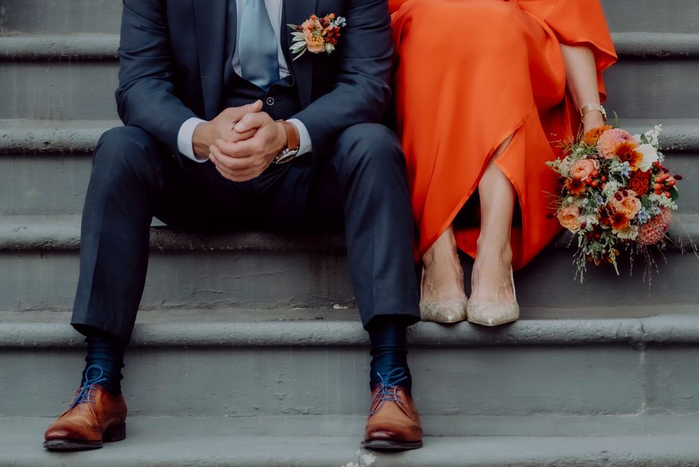 Bride in red dress and groom in navy suit sitting on steps with bouquet.