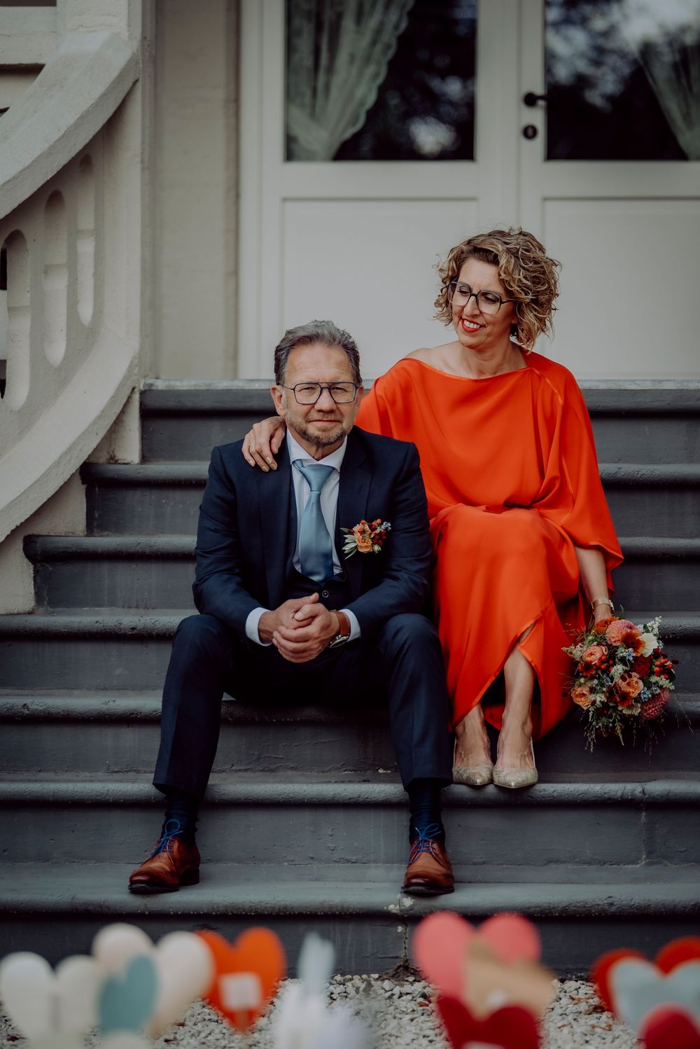 Smiling couple sitting on steps, bride in red dress holding colorful bouquet.