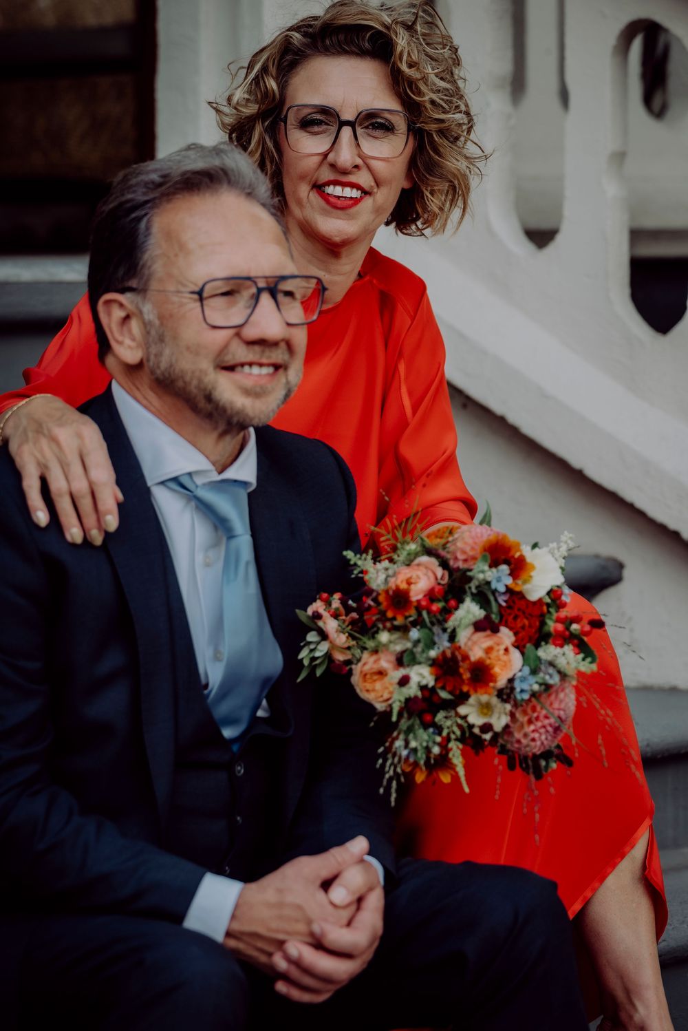 Happy couple posing on steps, woman in red dress holding vibrant bouquet beside smiling man.