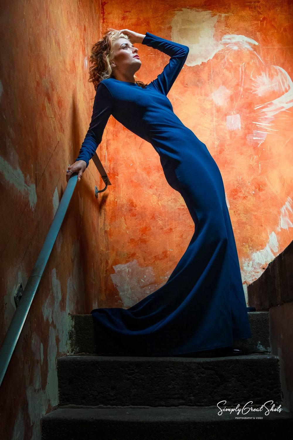 A figure in a flowing royal blue evening gown poses dramatically on dark stairs against a textured orange wall for Fashion Photography.