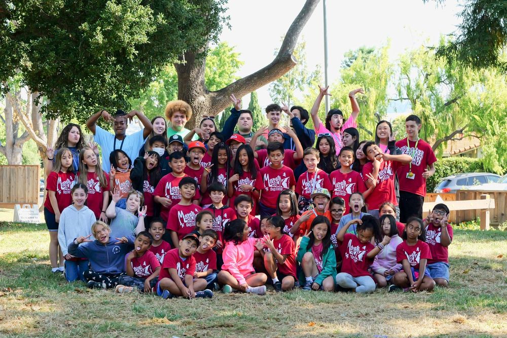 Large group of students sitting together outdoors under shady trees during a school event.