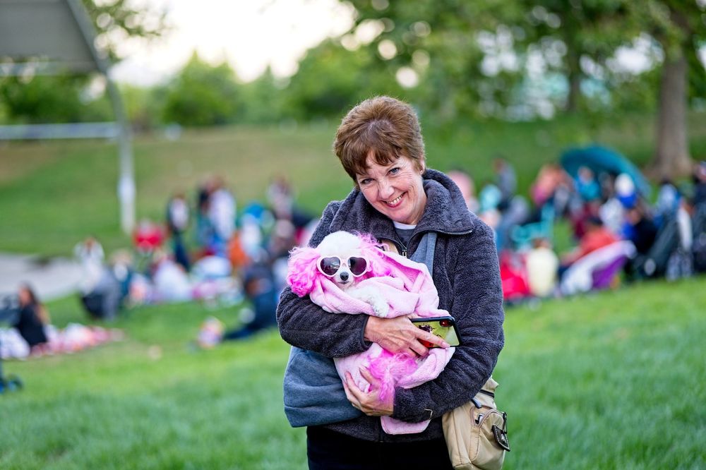 A person holds a pink plush toy while standing on a grassy hill with people in the background.
