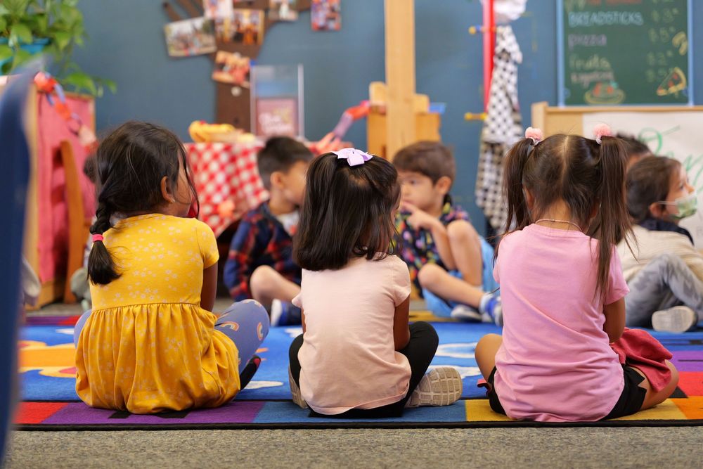 Children sit in colorful clothing on mats during a classroom activity.