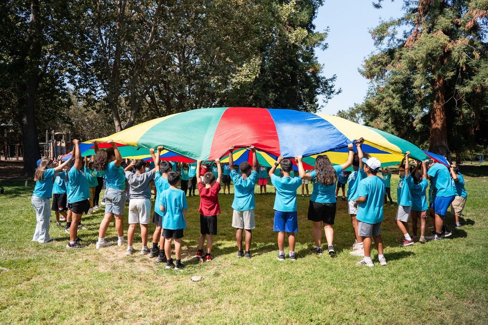 A group of children in blue shirts hold a colorful parachute together during an outdoor activity.