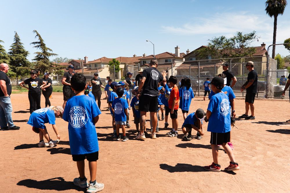 Children in blue shirts gather on a dirt field for outdoor activities with adult supervisors.