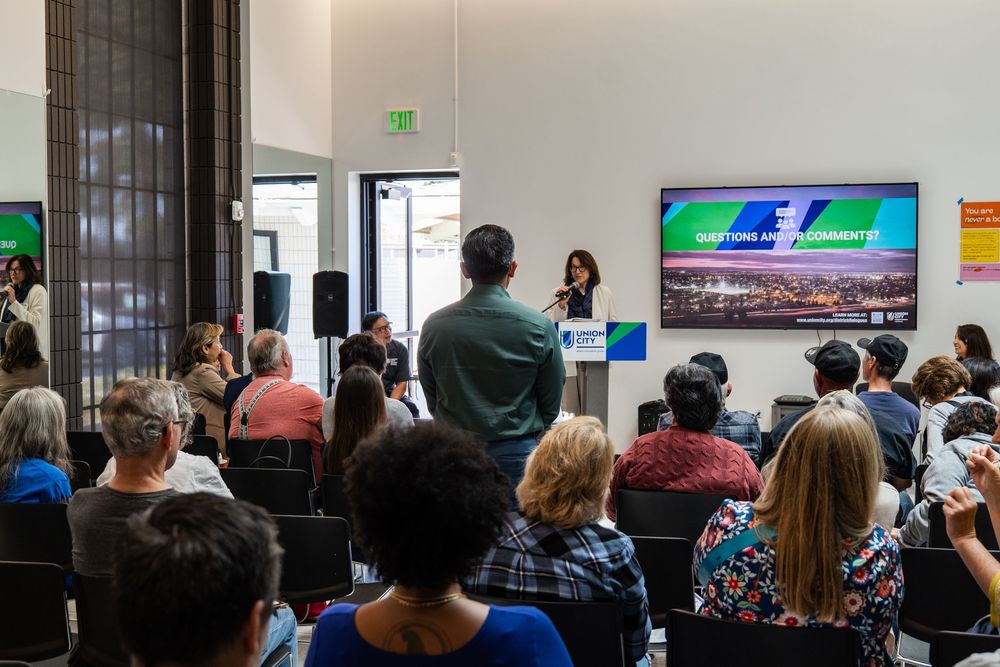 A speaker presents to a packed audience in a modern conference room setting.
