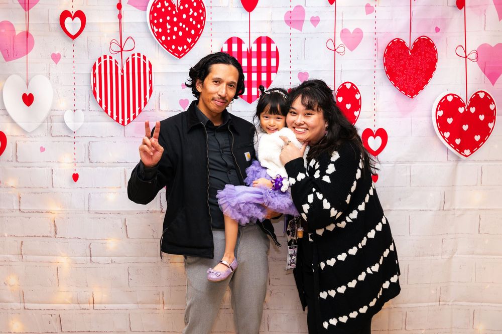 A couple poses with gifts against a pink heart-patterned backdrop during a celebration.