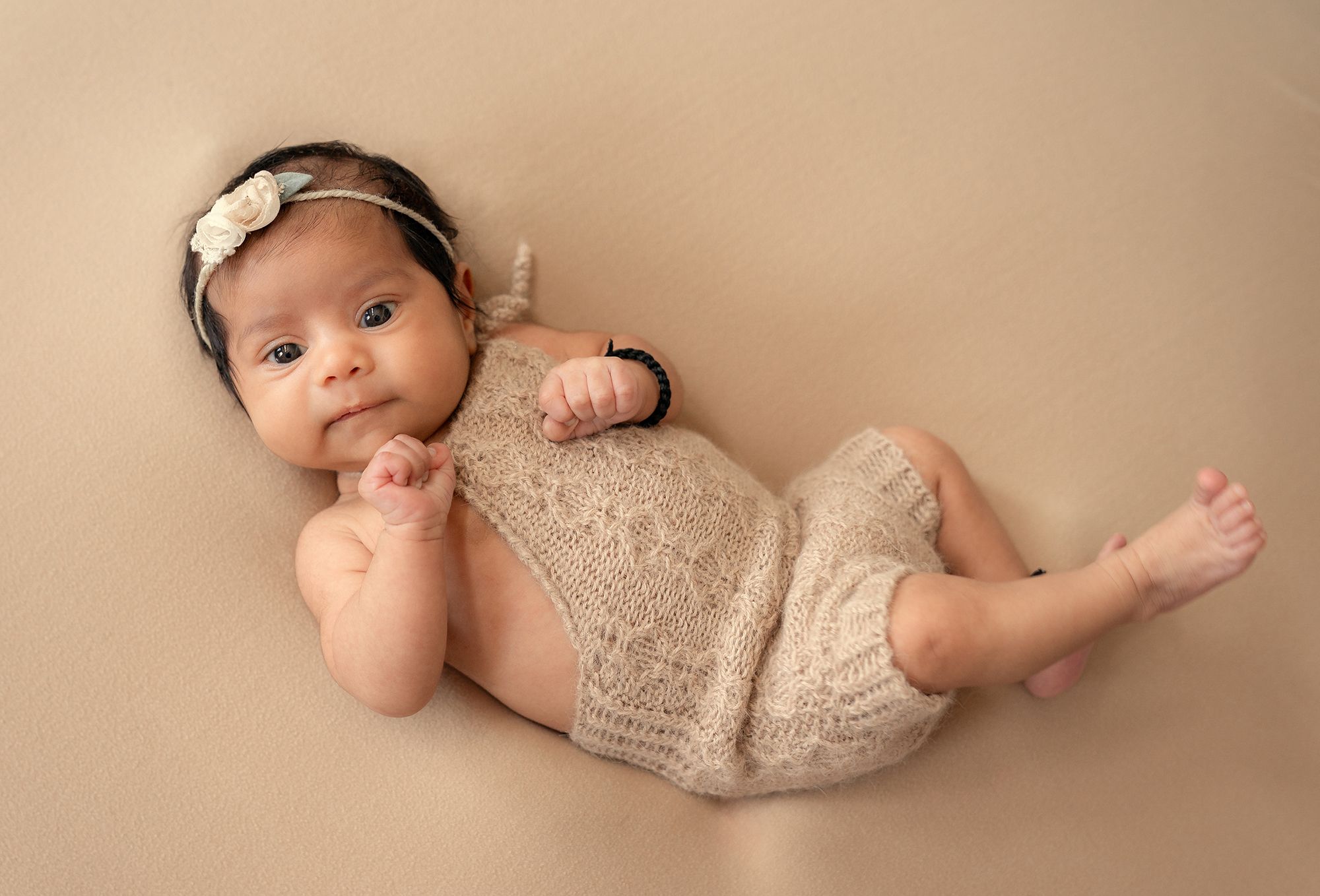 A newborn baby wearing a delicate beige romper lies peacefully on a soft beige backdrop during a professional photoshoot.
