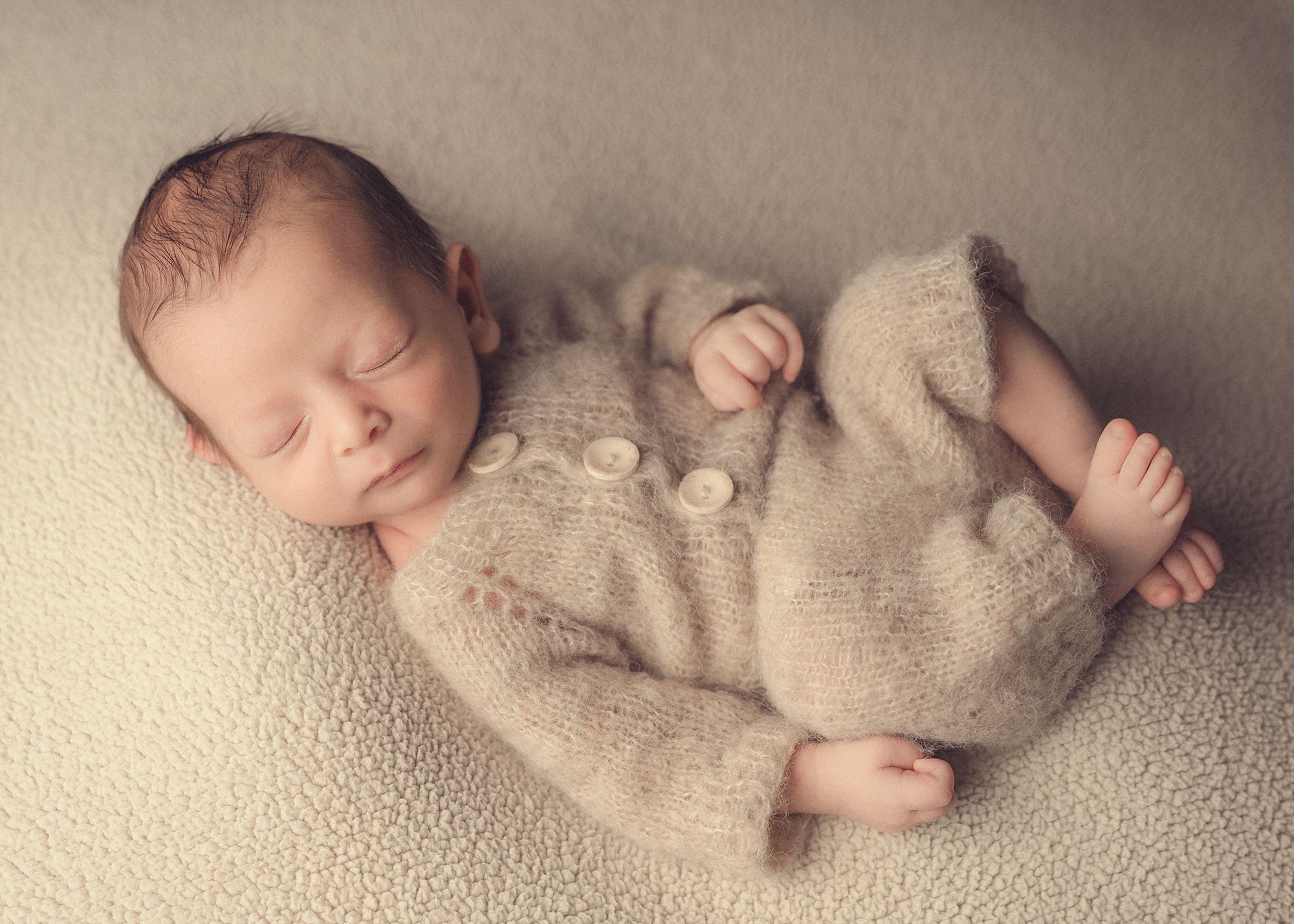 A sleeping newborn baby wearing a cozy knit onesie lies peacefully on a textured cream-colored blanket.