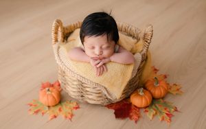 A sleeping newborn posed in an autumn themed setting with small pumpkins and fall leaves on a wooden floor.