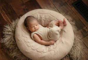 A sleeping newborn baby curled up on a round cream-colored knitted cushion in warm natural lighting.