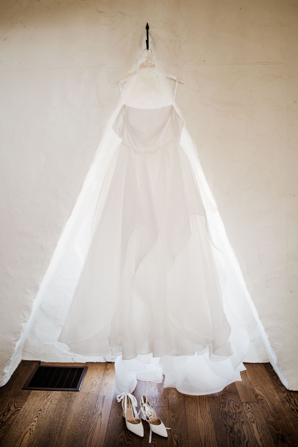 Ethereal white wedding gown hanging against neutral wall with shoes beneath.