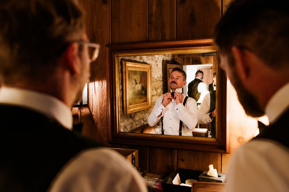Groomsmen getting ready in a rustic room with wood paneled walls and framed artwork.