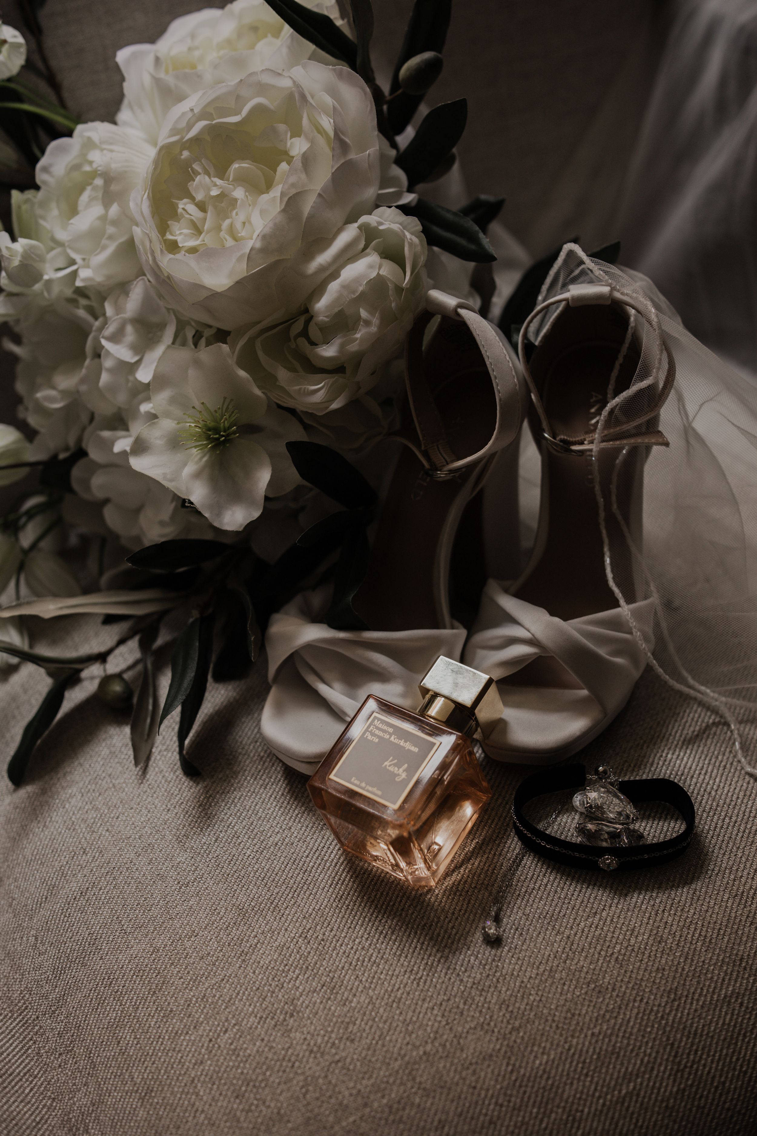 Elegant wedding flat lay featuring white peony bouquet, nude heels, perfume bottle, and jewelry on linen.