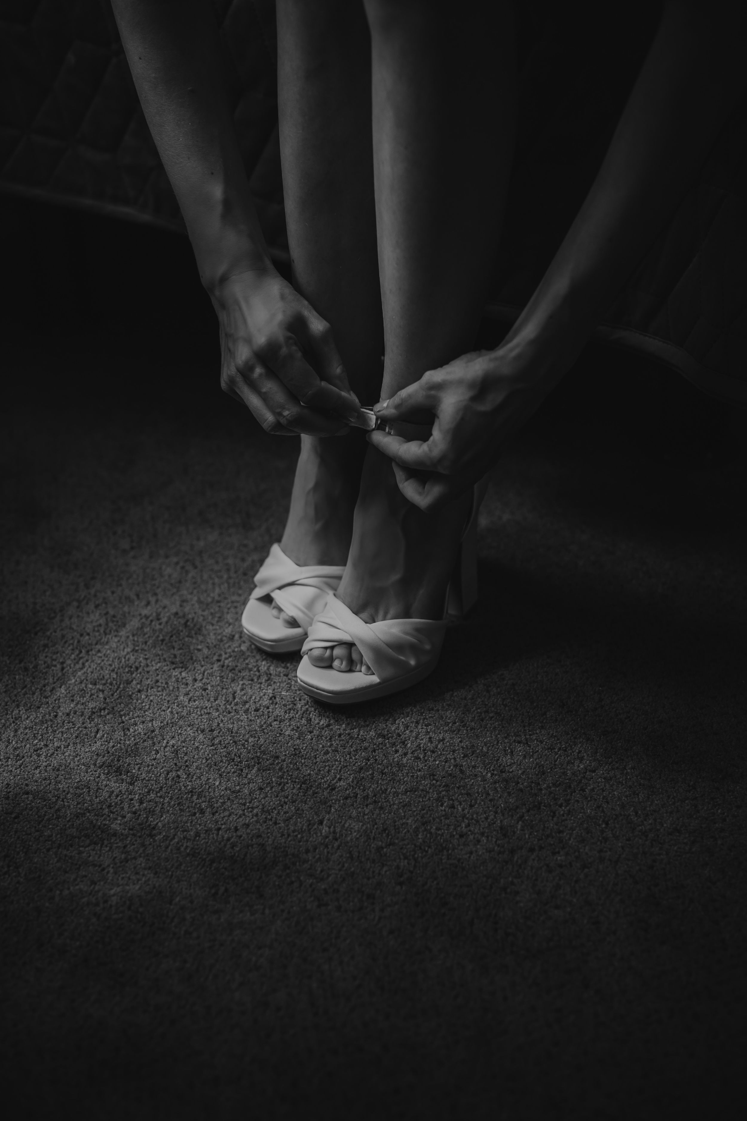 Black and white photo of a bride fastening strappy heeled sandals on her feet before her wedding.