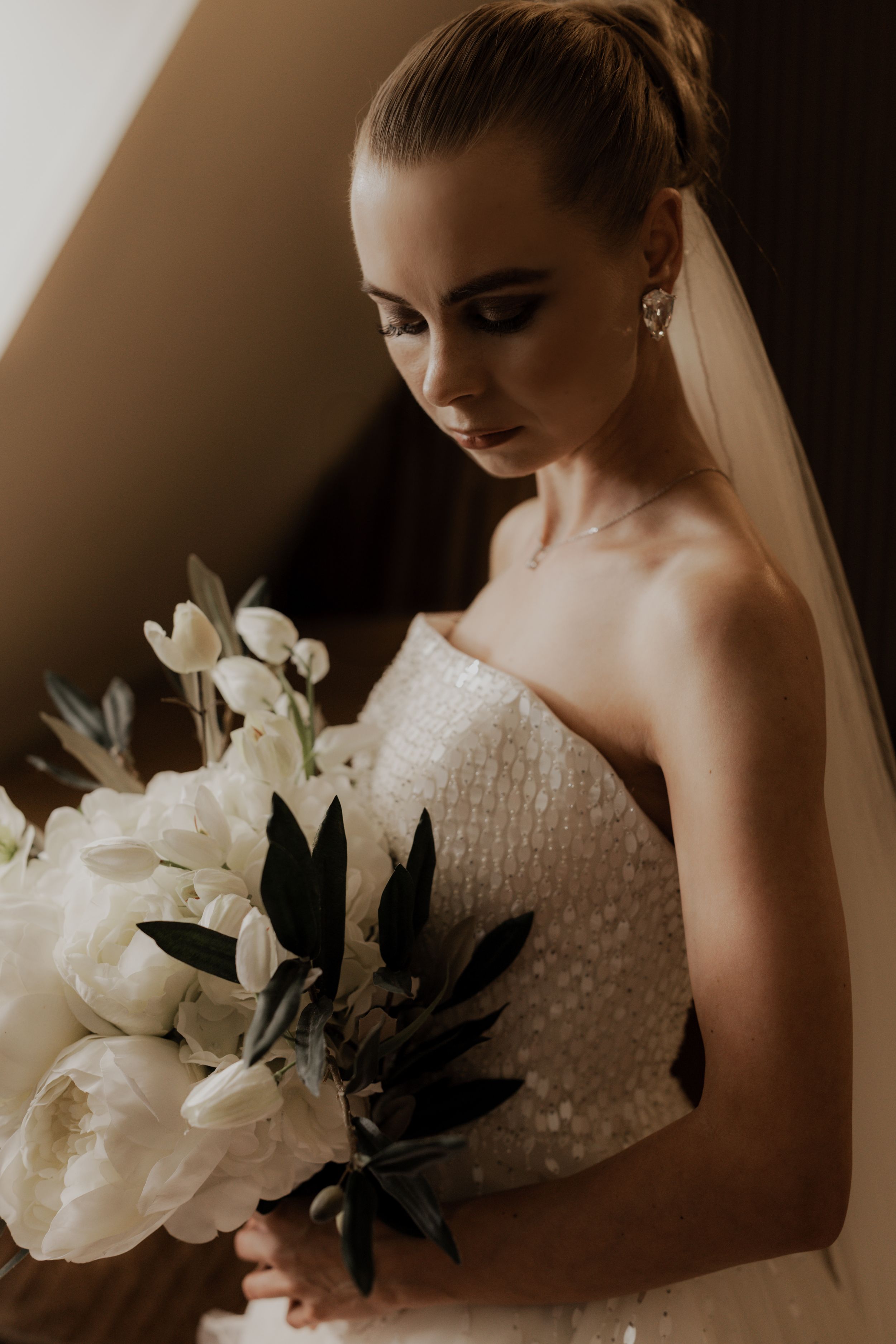 Elegant bride in strapless sequined gown holding white tulip and peony bouquet with dark foliage.