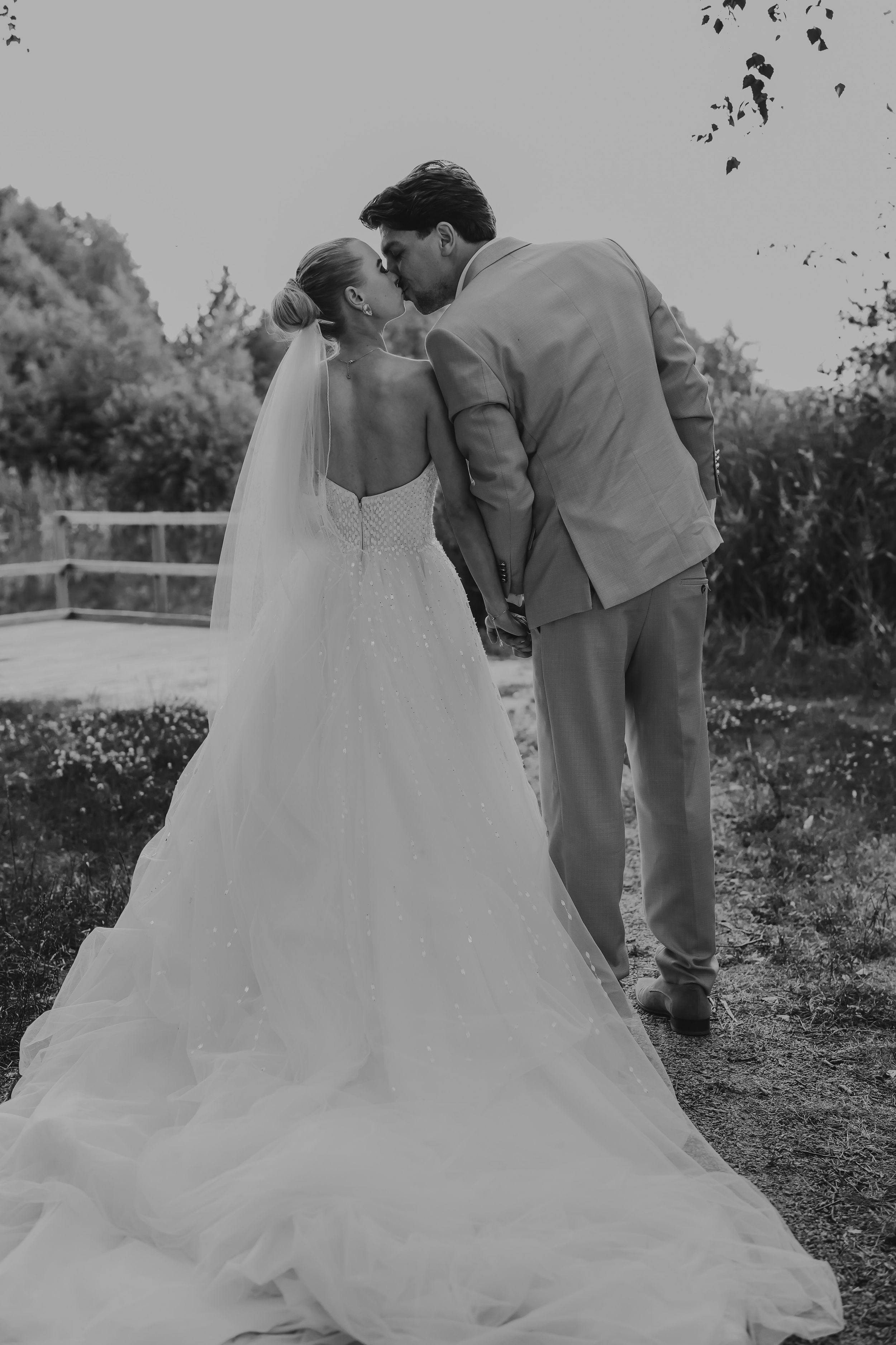 Black and white photo of a groom kissing his bride on the cheek in a garden setting.