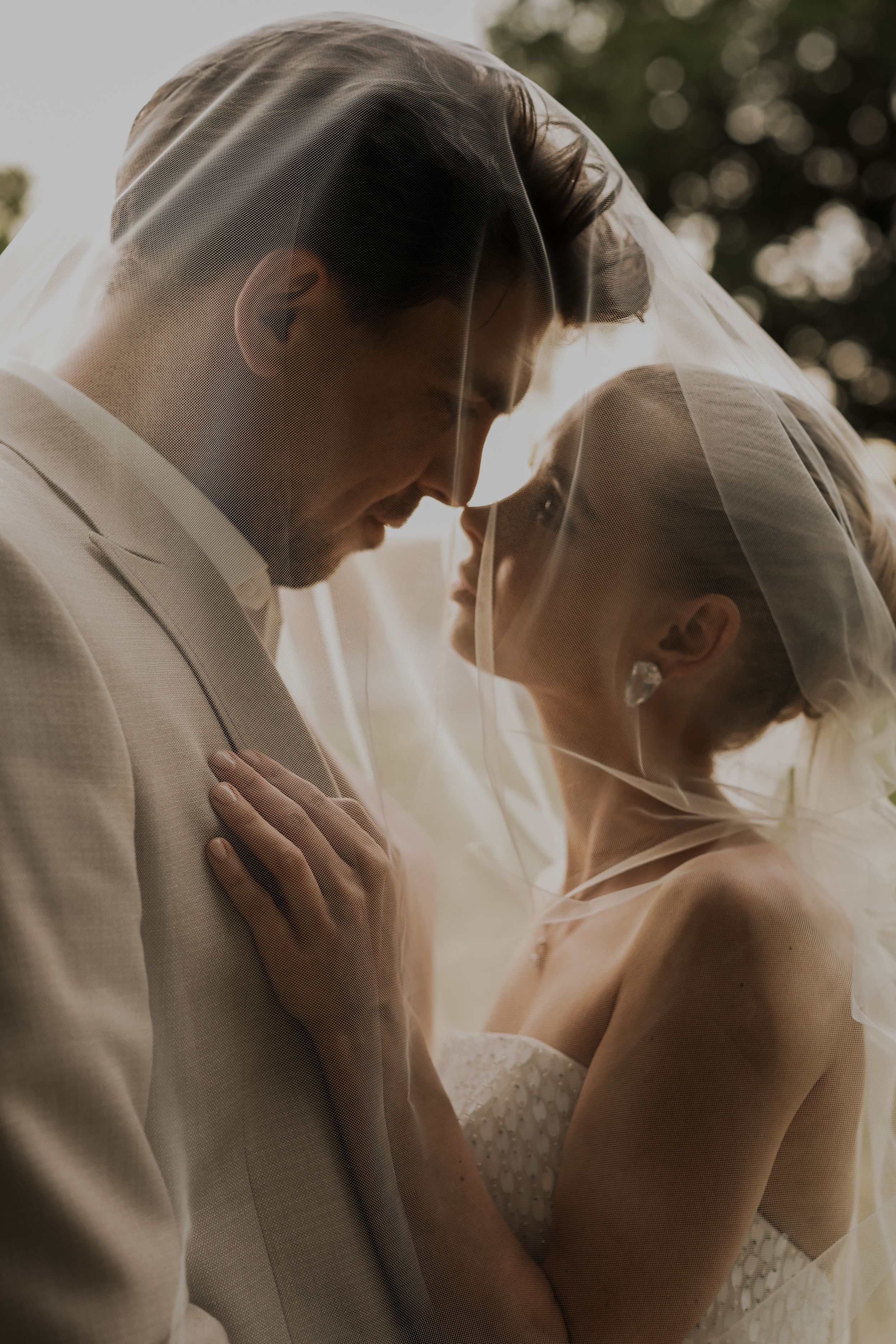 Bride and groom touching foreheads tenderly under a sheer veil in warm golden light.