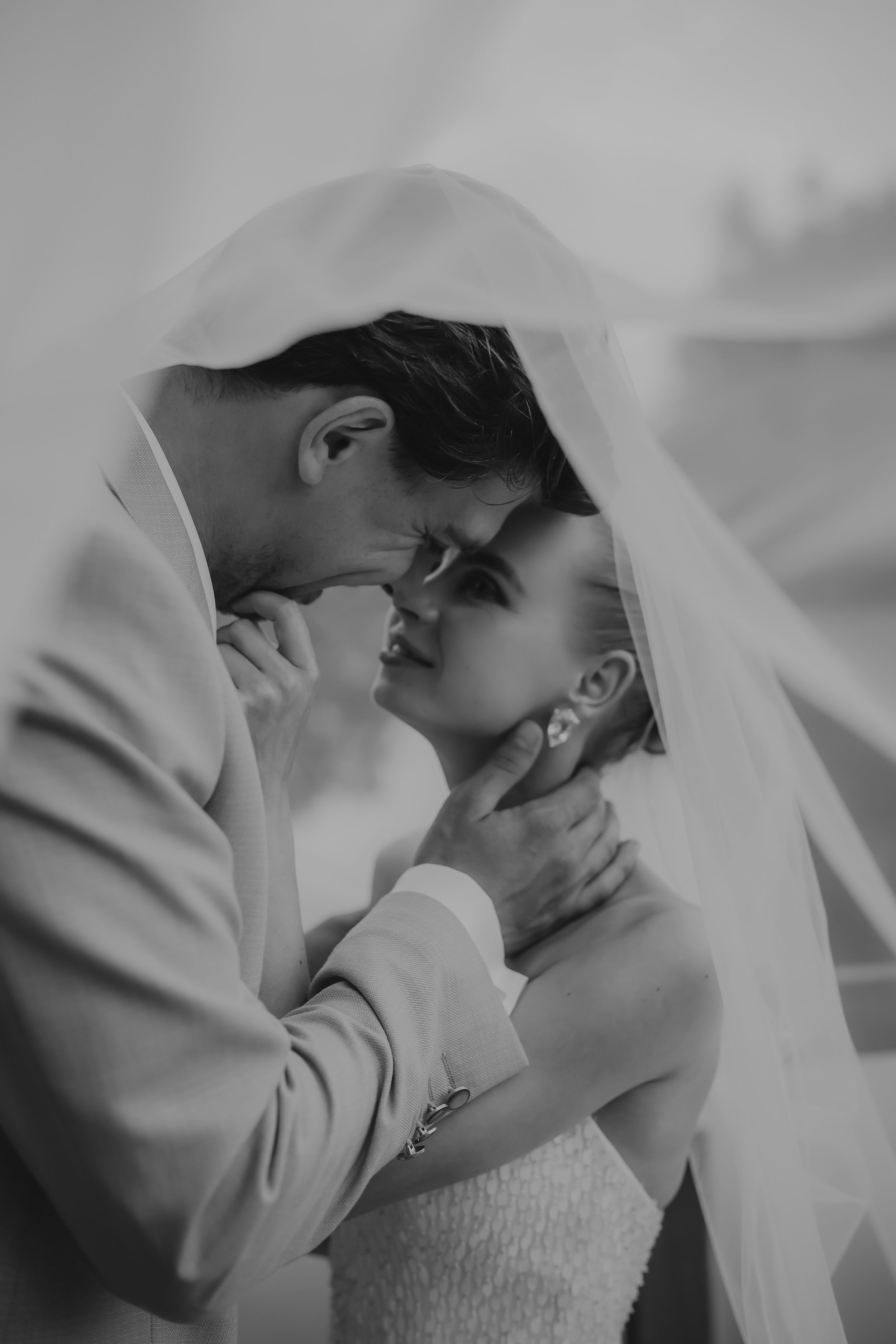Black and white romantic photo of bride and groom sharing intimate moment under flowing wedding veil.