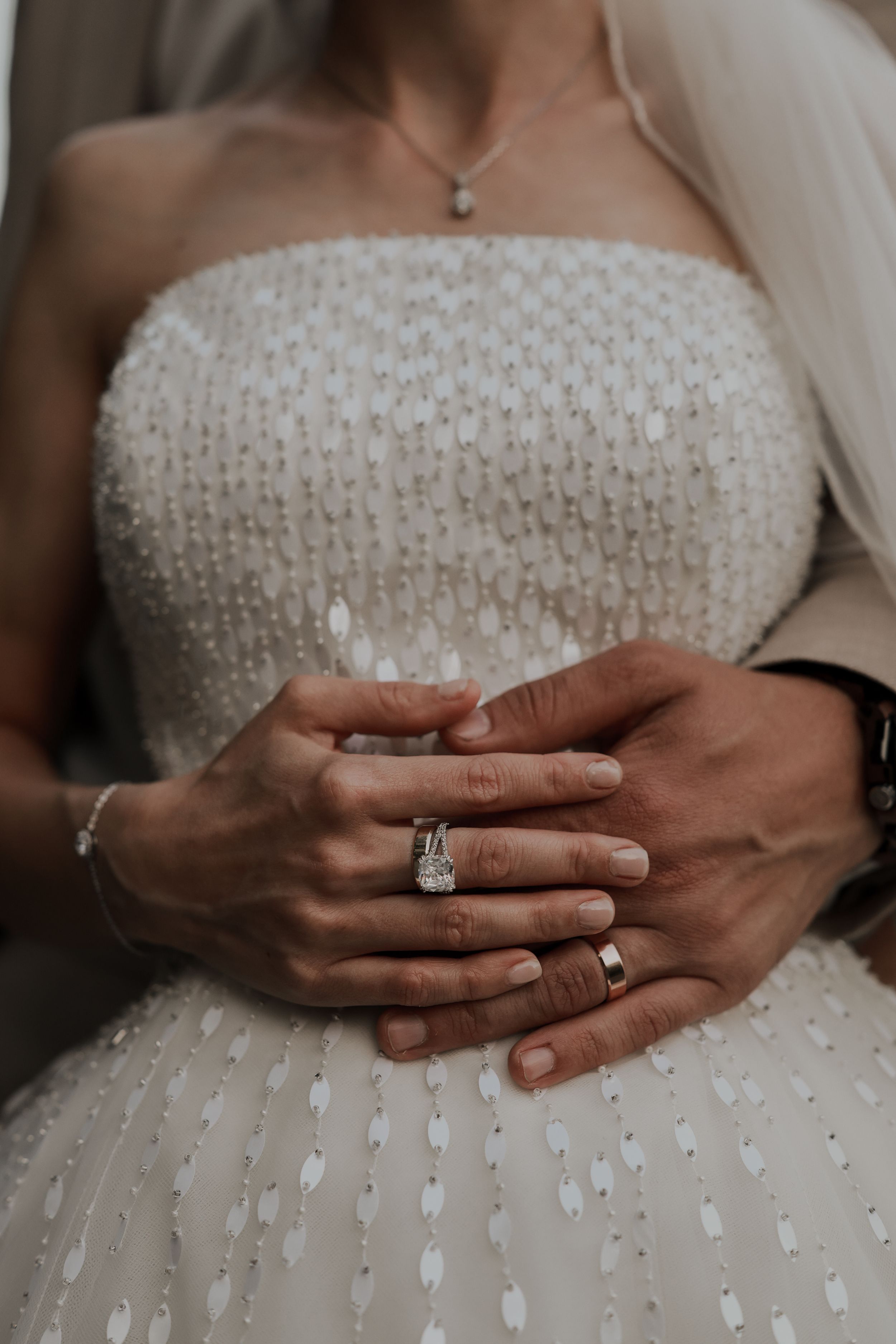 Close-up of bride and groom hands showing diamond engagement ring and wedding bands on white gown.