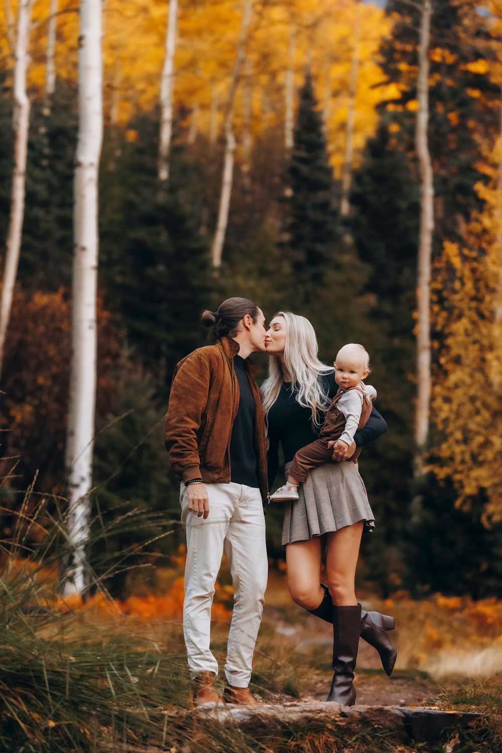 Couple kissing while holding baby in colorful autumn birch forest setting.