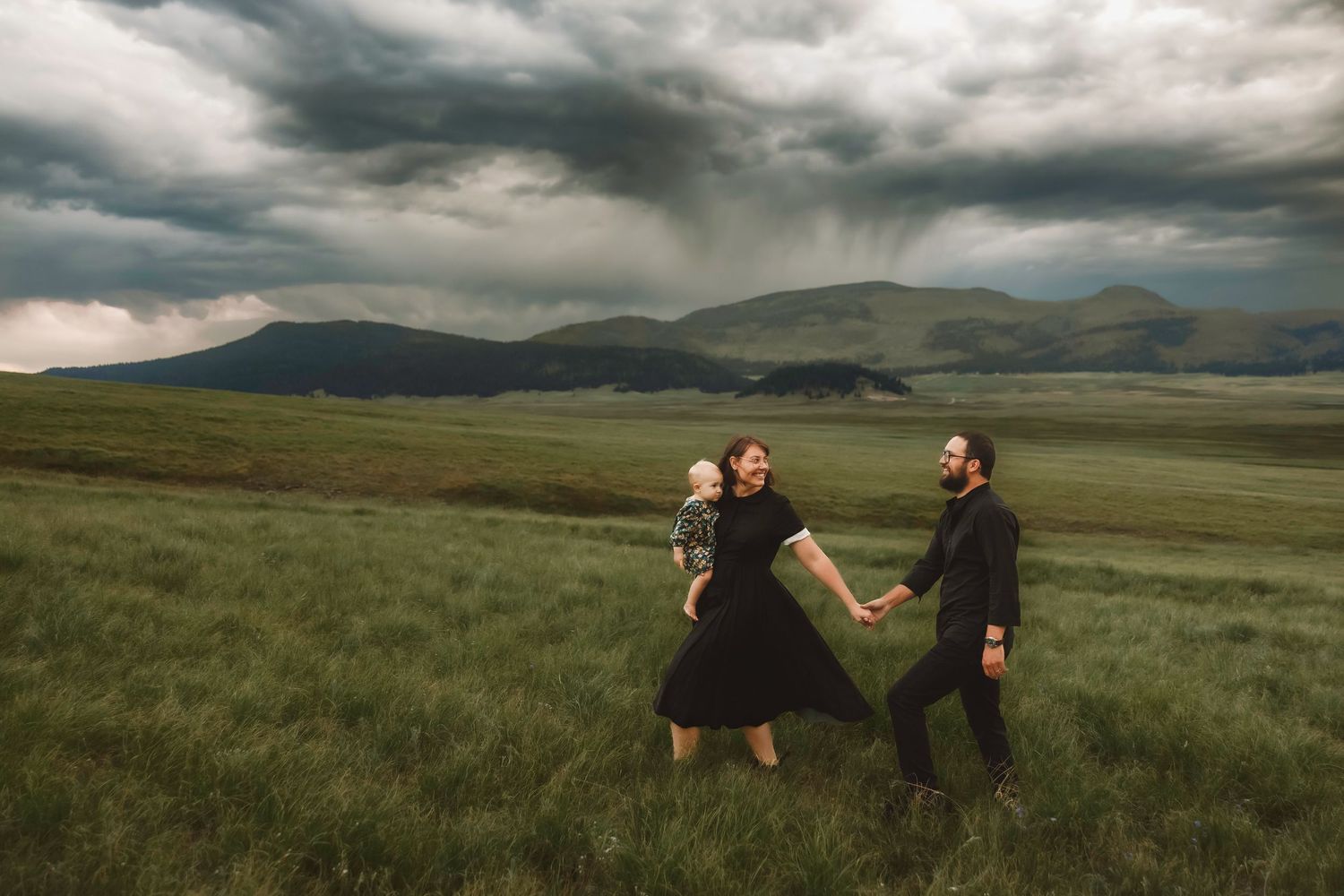 Couple in black outfits hold hands in vast green meadow under dramatic stormy sky with mountain backdrop.