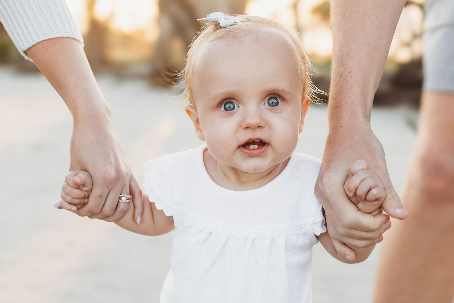 Toddler in white dress reaching toward camera during golden hour outdoor photoshoot.
