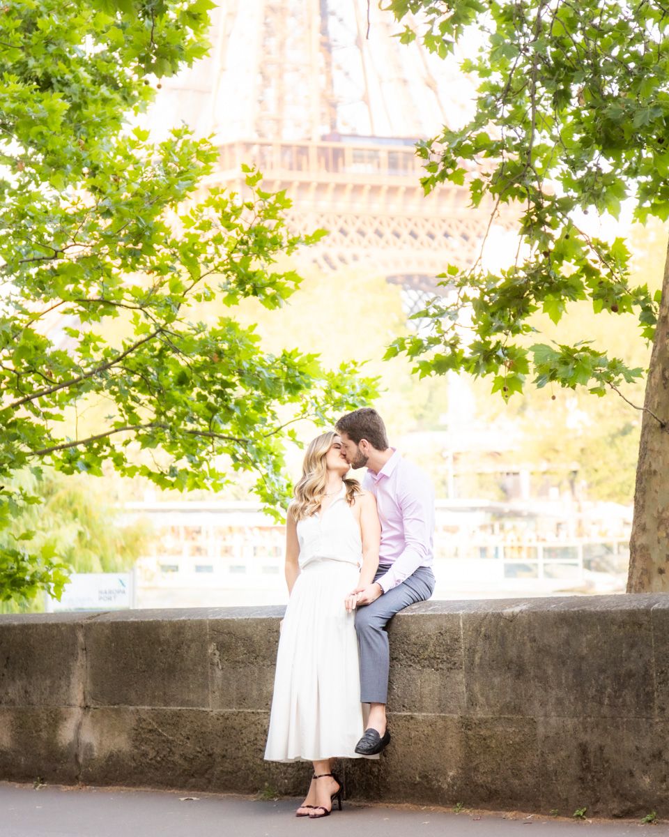 A romantic couple shares tender moments along a stone wall beneath green trees in a park-like setting.
