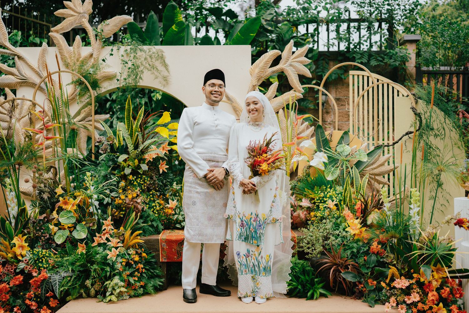 Couple in traditional Malay wedding attire posing before a lush tropical floral backdrop with vibrant orange and yellow blooms.