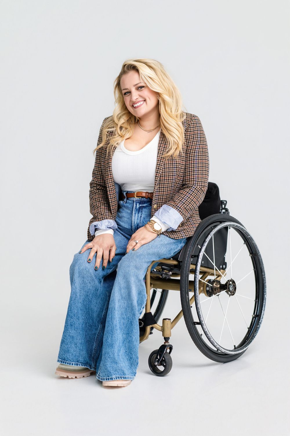 Stylish woman in plaid blazer and jeans sitting confidently in wheelchair against white background.