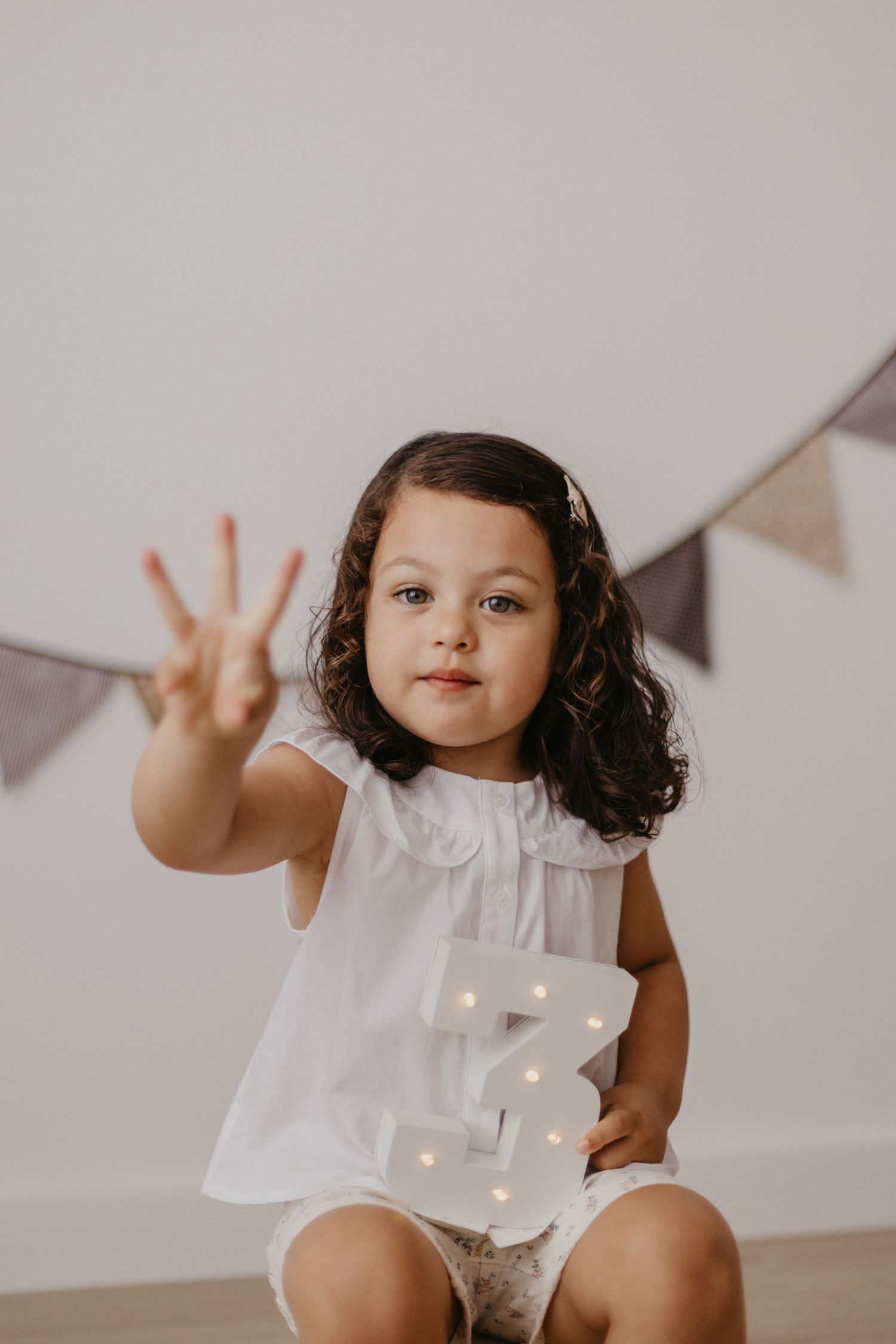 Child in white outfit making playful gestures against wall with decorative bunting.
