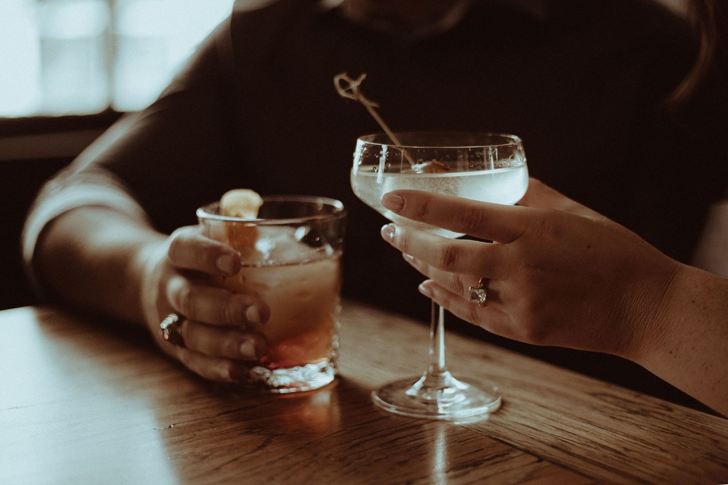 Two cocktail glasses clink together in dim atmospheric lighting.