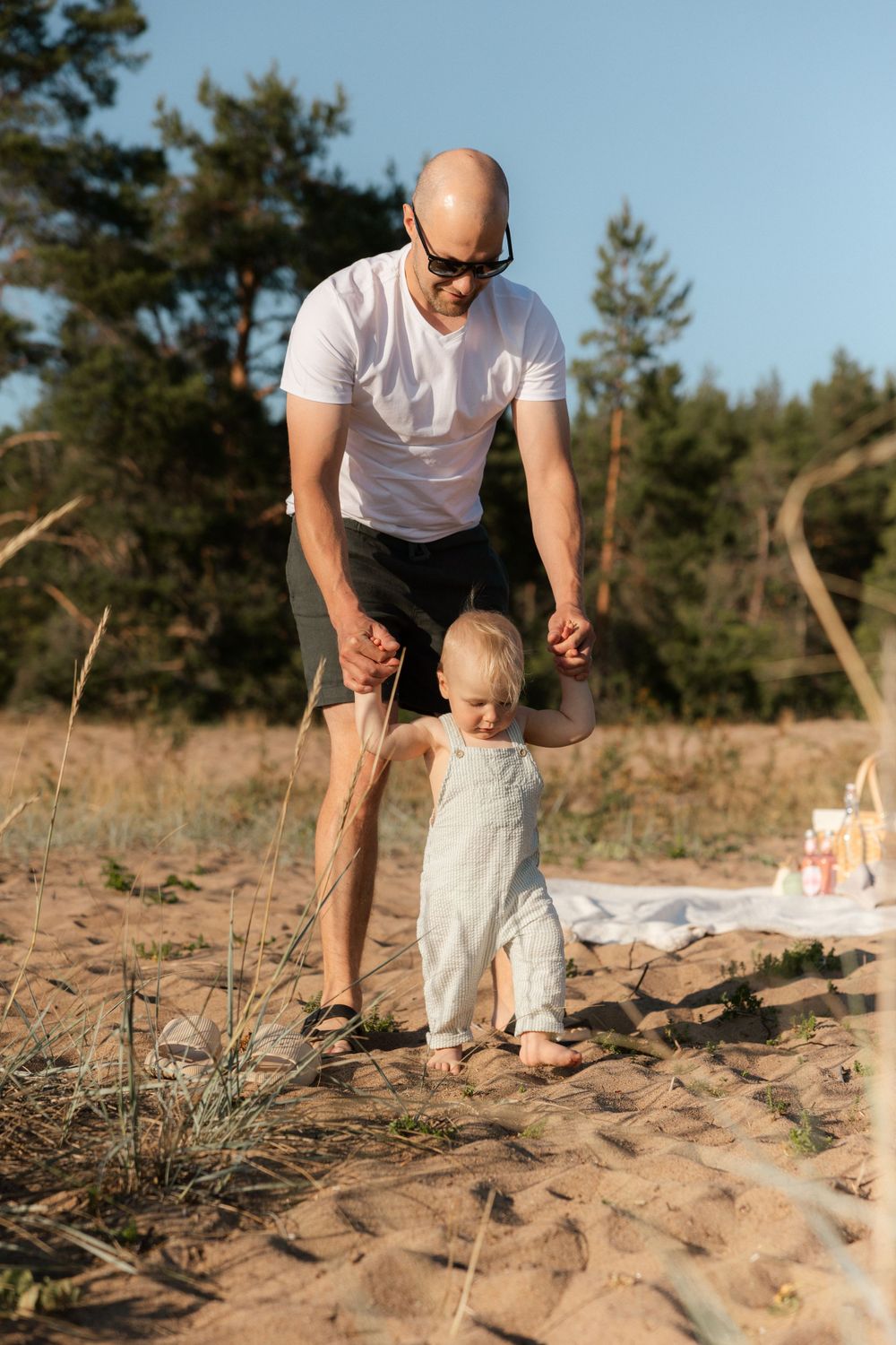 Adult in white t-shirt helps small toddler take first steps on sandy beach with pine trees in background.