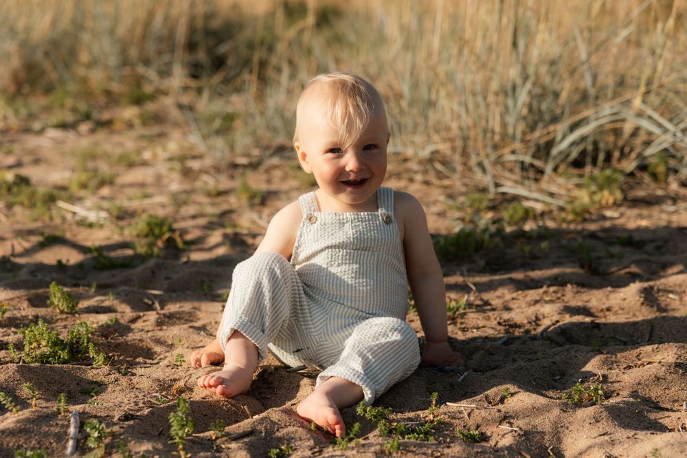 A small child in white overalls sits barefoot on sandy ground during golden hour.