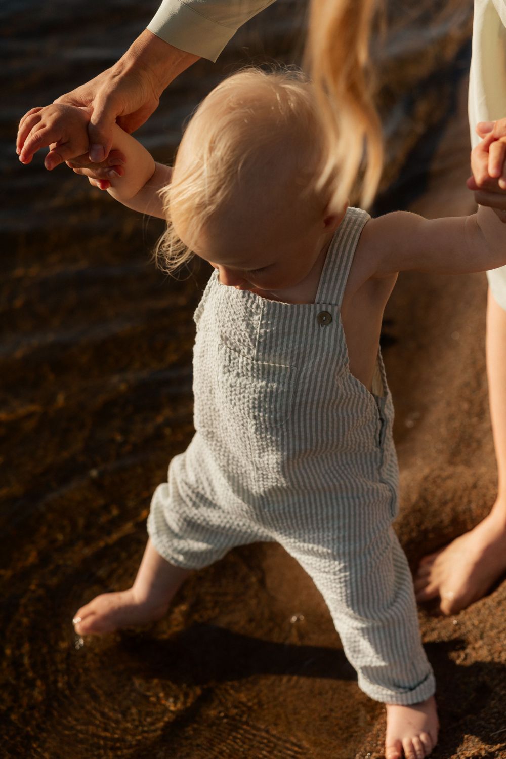 A small child in striped overalls takes early steps while being supported by adult hands at sunset.