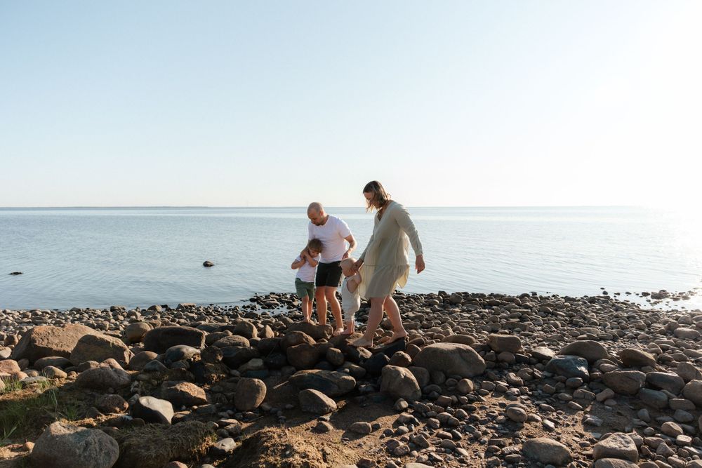 Two people walk together on a rocky shoreline beside calm waters during a sunny day.