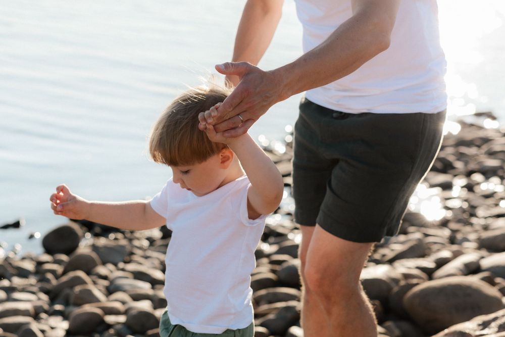 A child in a white shirt stands on a rocky beach while someone in shorts gently touches their head.