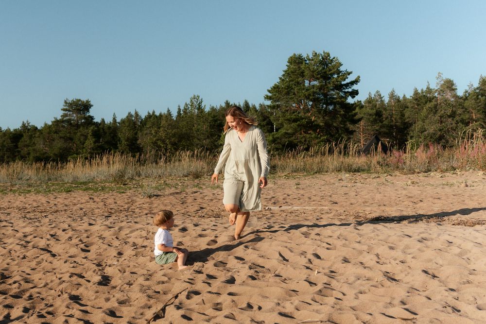 Two people walking together on a sandy beach near pine trees on a sunny day.