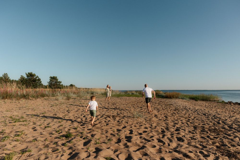 People walking along a sandy beach with tall grass and ocean views on a bright sunny day.