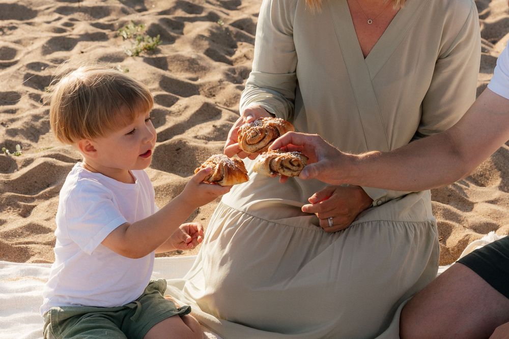 A small child and adult sit together on a sandy beach sharing food at sunset.