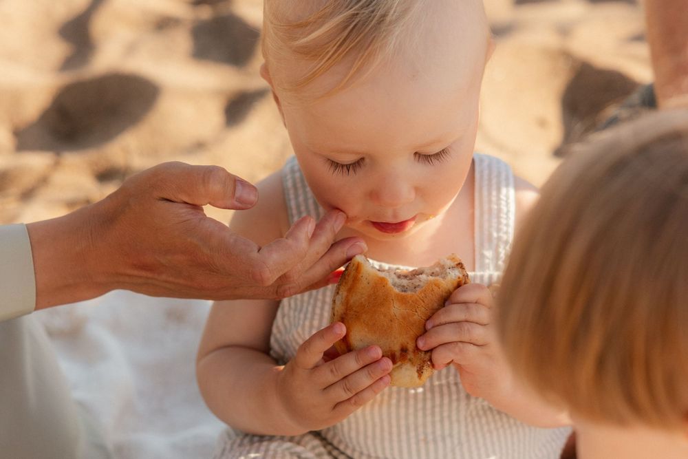 Small child in striped outfit takes a bite of pastry while being helped by adult hands in sunny outdoor setting.