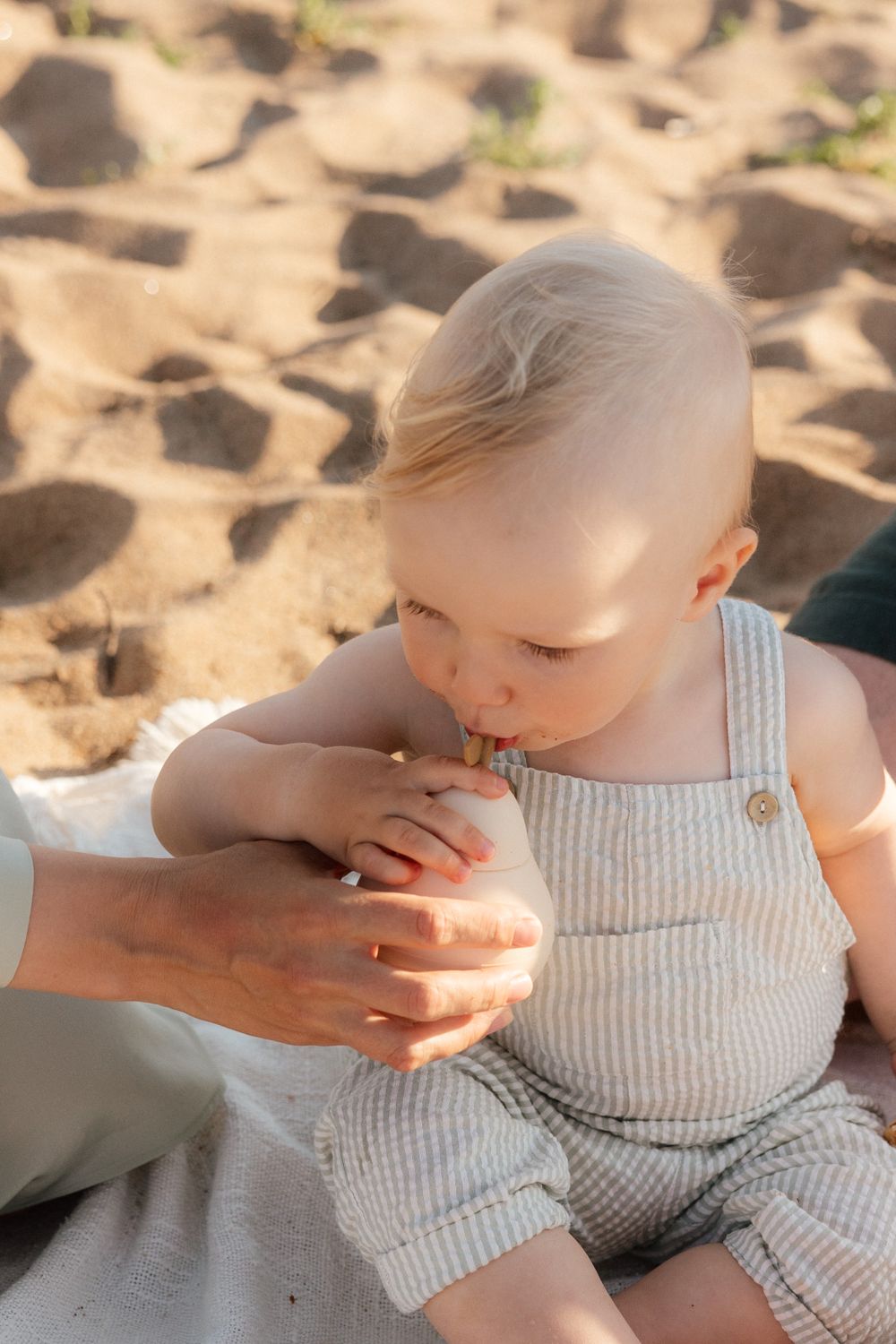 Young child in white overalls sits on sandy beach while being comforted by gentle hands.