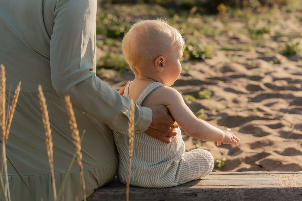 A parent sits with their small baby in striped overalls together on the sand at sunset.